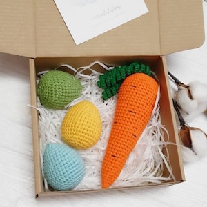 Puede incluir: Una caja de regalo llena de papel de seda blanco y decoraciones de Pascua de ganchillo. Las decoraciones incluyen tres huevos en verde, amarillo y azul, y una zanahoria con hojas verdes.