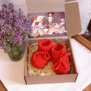 May include: A pair of red knitted baby booties with a bow on each one, inside a brown cardboard box with shredded paper.