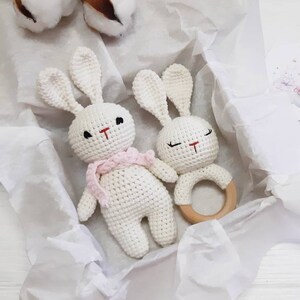 Puede incluir: Dos juguetes de conejo de crochet blanco con ojos negros y narices rojas. Un conejo lleva una bufanda rosa. Los juguetes de conejo est&aacute;n en una caja de regalo blanca con papel de seda blanco. Un anillo de dentici&oacute;n de madera est&aacute; al lado de los juguetes de conejo.