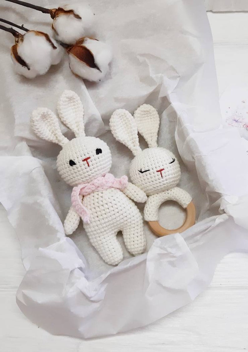 Puede incluir: Dos juguetes de conejo de crochet blanco con ojos negros y narices rojas. Un conejo lleva una bufanda rosa. Los juguetes de conejo est&aacute;n en una caja de regalo blanca con papel de seda blanco. Un anillo de dentici&oacute;n de madera est&aacute; al lado de los juguetes de conejo.