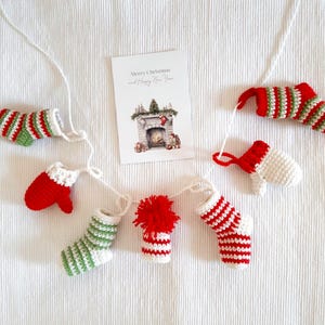 Può includere: Una ghirlanda natalizia fatta a mano con calzini e guanti all'uncinetto in miniatura rossi, bianchi e verdi. È presente anche una cartolina con un'illustrazione di un camino e il testo "Merry Christmas and Happy New Year".