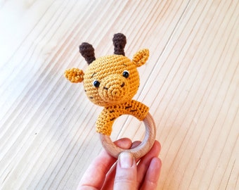 May include: A crocheted giraffe toy with brown ears and a wooden ring. The giraffe is yellow and has black eyes.