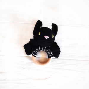 May include: A black crochet bat-shaped baby rattle with a wooden ring. The bat has yellow eyes and a pink nose. The wooden ring has white text. The rattle is on a white wooden surface.