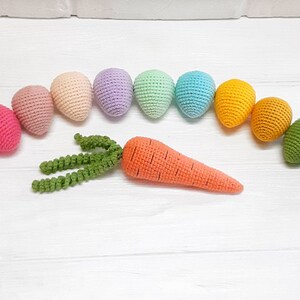 Puede incluir: Un surtido de huevos de crochet arco&iacute;ris con una zanahoria y hojas verdes de crochet. Los huevos est&aacute;n en orden de arco&iacute;ris, comenzando con rosa y terminando con azul.