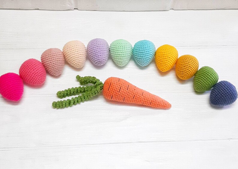 Puede incluir: Un surtido de huevos de crochet arco&iacute;ris con una zanahoria y hojas verdes de crochet. Los huevos est&aacute;n en orden de arco&iacute;ris, comenzando con rosa y terminando con azul.