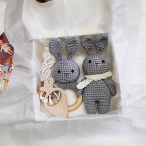 Puede incluir: Dos juguetes de conejo de crochet gris con anillos de dentici&oacute;n de madera y una bufanda de crochet blanco en un conejo. Los juguetes est&aacute;n en una caja de regalo blanca con papel de seda blanco.