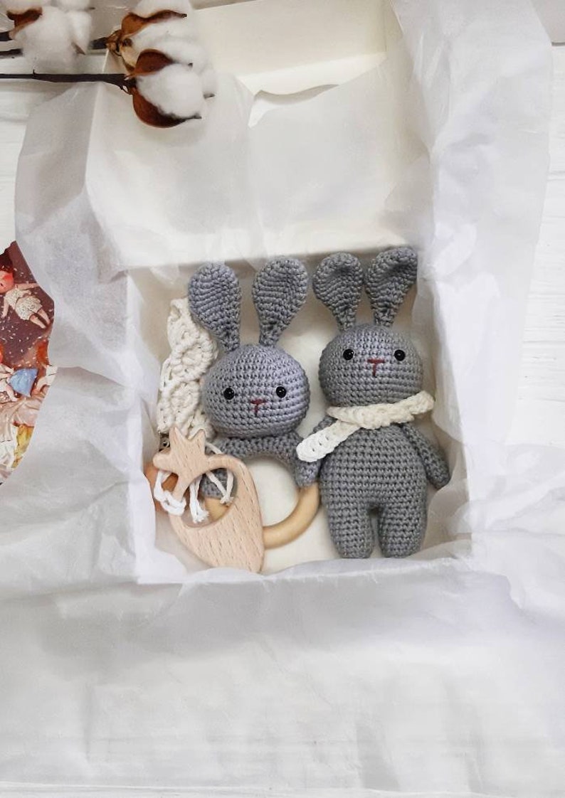 Puede incluir: Dos juguetes de conejo de crochet gris con anillos de dentici&oacute;n de madera y una bufanda de crochet blanco en un conejo. Los juguetes est&aacute;n en una caja de regalo blanca con papel de seda blanco.