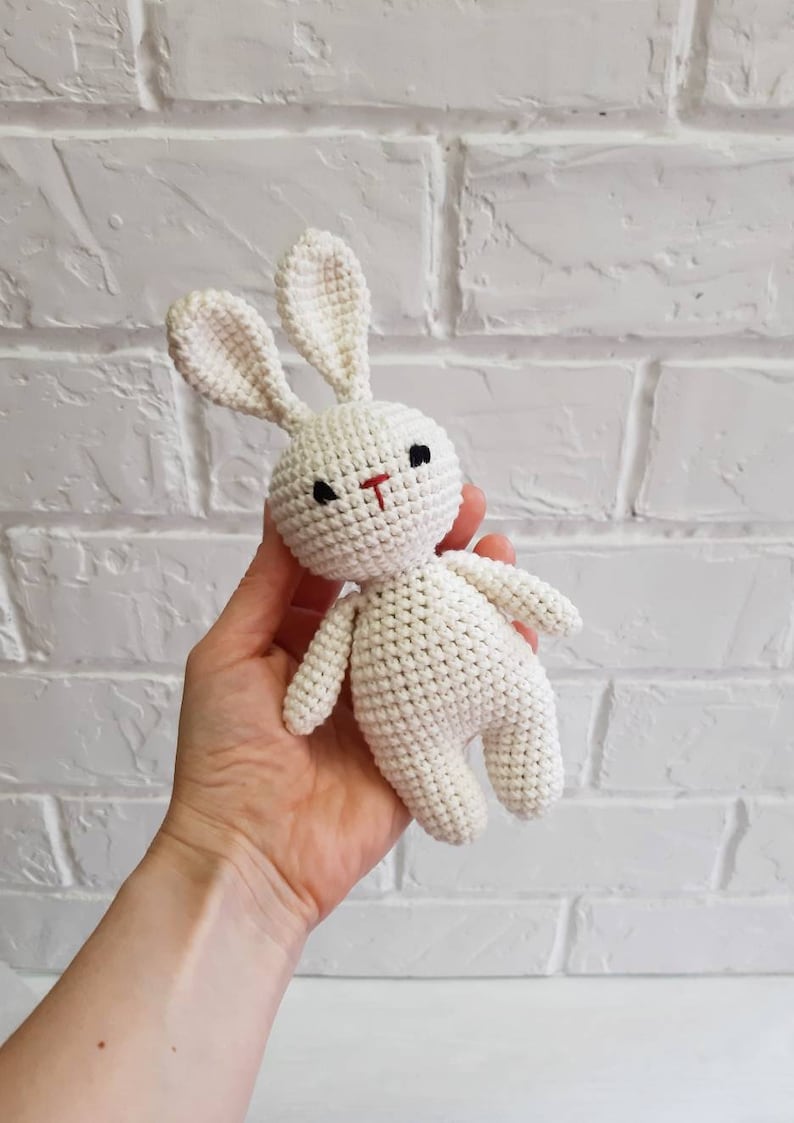 Puede incluir: Un juguete de conejo blanco de crochet con ojos negros y nariz roja. El conejo est&aacute; siendo sostenido en la mano de una persona.