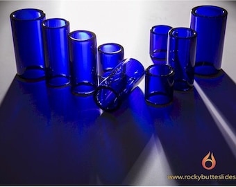 May include: A set of eight blue glass guitar slides of varying sizes. The slides are arranged in a semi-circle on a white surface.