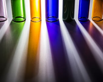 May include: A row of six glass bottles in different colors, including purple, green, orange, blue, and clear. The bottles are lined up on a white surface, casting colorful shadows.