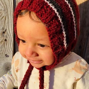 May include: A burgundy crocheted baby bonnet with white and black stripes. The bonnet has long ties and is worn by a baby. The baby is wearing a white long-sleeved shirt with a pattern of bears.