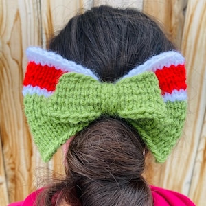 Può includere: Una fascia per capelli all'uncinetto verde, rossa e bianca con un design a fiocco. La fascia è verde con un fiocco al centro e dettagli rossi e bianchi sui lati. La fascia è indossata sulla testa di una persona.