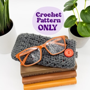 May include: A crochet pattern for a gray headband with a pair of orange glasses and a red button on top of a stack of books. The books are titled "The Return of the King" and "The Two Towers". The text "@greenforfarms" is written on a wooden sign.