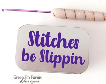 May include: A silver tin with the text "Stitches be Slippin'" in purple lettering. The tin is sitting on a white surface next to a pink and white crochet hook.