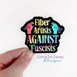 May include: A black and iridescent sticker with the text "Fiber Artists Against Fascists" and a raised fist holding a knitting needle and a pair of scissors. The sticker is on a white background.