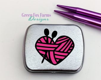 May include: A silver metal tin with a pink and black heart design featuring a ball of yarn and knitting needles. The text "Green Fox Farms Designs" is printed on the tin.