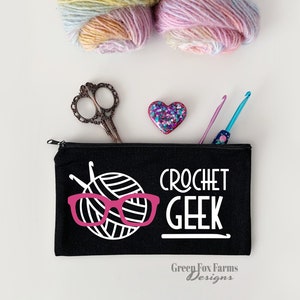 May include: Black zippered pouch with the text "Crochet Geek" in white lettering. A white yarn ball with pink glasses is pictured on the pouch. The pouch is on a white surface with yarn balls, scissors, and a crochet hook.