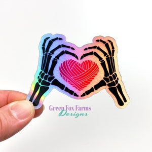 May include: A holographic sticker featuring two black skeleton hands forming a heart shape around a red yarn ball heart. The text Green Fox Farms Designs is printed in purple and teal below the skeleton hands on the sticker surface.
