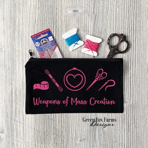 May include: Black zippered pouch with pink embroidery of sewing tools and the text "Weapons of Mass Creation". The pouch is on a woodgrain surface with two spools of thread and a pair of scissors.