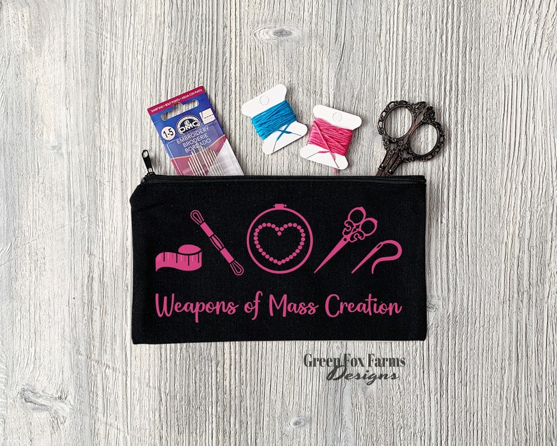 May include: Black zippered pouch with pink embroidery of sewing tools and the text "Weapons of Mass Creation". The pouch is on a woodgrain surface with two spools of thread and a pair of scissors.