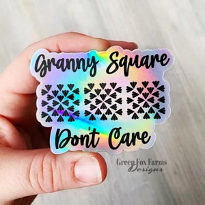 May include: A holographic sticker with the text "Granny Square Don't Care" and a design of three granny squares. The sticker has a rainbow iridescent background and black text. The text "Green Fox Farms Designs" is at the bottom of the sticker.