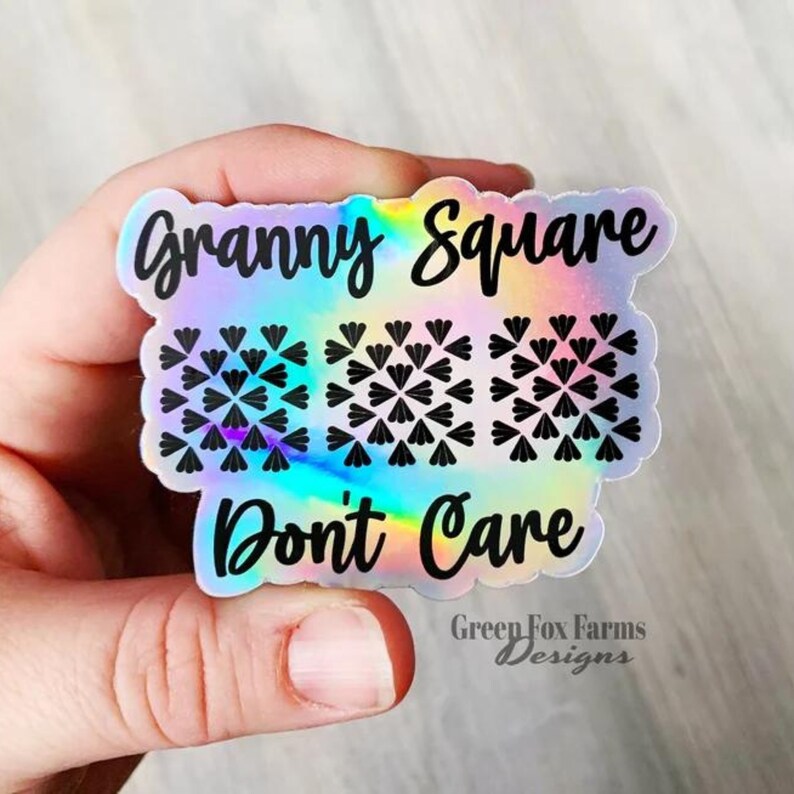 May include: A holographic sticker with the text "Granny Square Don't Care" and a design of three granny squares. The sticker has a rainbow iridescent background and black text. The text "Green Fox Farms Designs" is at the bottom of the sticker.