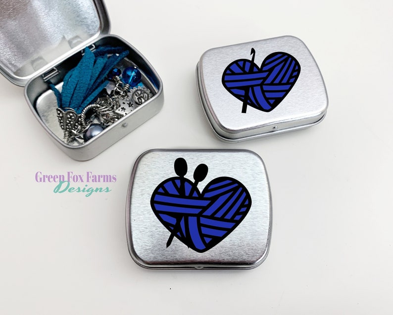 Puede incluir: Tres latas de metal plateadas con un dise&ntilde;o en forma de coraz&oacute;n que presenta hilo azul y agujas de tejer. El texto "Green Fox Farms Designs" es visible en una de las latas.