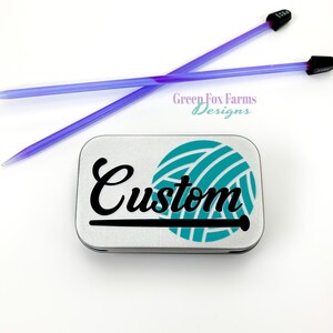 May include: A silver metal tin with a teal yarn ball and black lettering that says "Custom". The tin is for storing knitting needles. Two purple knitting needles are in the background.