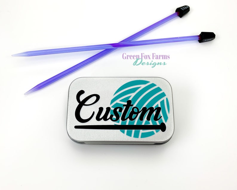 May include: A silver metal tin with a teal yarn ball and black lettering that says "Custom". The tin is for storing knitting needles. Two purple knitting needles are in the background.