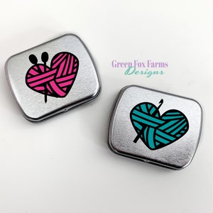 Puede incluir: Dos latas de metal plateadas con un dise&ntilde;o en forma de coraz&oacute;n. El coraz&oacute;n de la lata de la izquierda es rosa con rayas negras y el coraz&oacute;n de la lata de la derecha es turquesa con rayas negras. Ambos corazones tienen una aguja de tejer negra y lana en su interior.