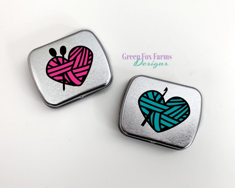 Puede incluir: Dos latas de metal plateadas con un dise&ntilde;o en forma de coraz&oacute;n. El coraz&oacute;n de la lata de la izquierda es rosa con rayas negras y el coraz&oacute;n de la lata de la derecha es turquesa con rayas negras. Ambos corazones tienen una aguja de tejer negra y lana en su interior.