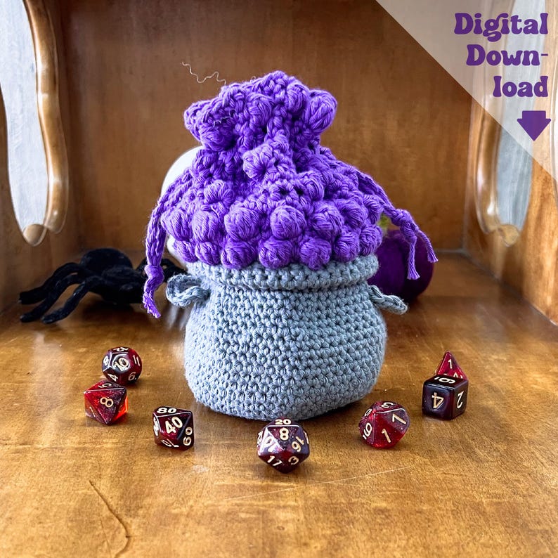 May include: A handmade crochet dice bag in gray and purple. The bag has a textured, rounded shape with a drawstring closure. Several polyhedral dice in red and black are scattered around the bag. The text "Digital Download" is visible.