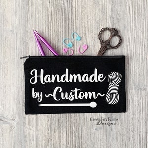 May include: Black zippered pouch with the text "Handmade by ~Custom~" in white, a white yarn ball, and a knitting needle. The pouch also has the text "Green Fox Farms Designs" in white.