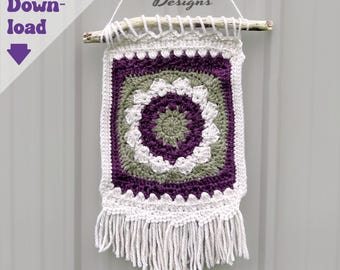 May include: A handmade, square crochet wall hanging with a central floral design in shades of purple, green, and white. The piece is suspended from a wooden dowel and features a fringe at the bottom. The text "Digital Download" is visible.