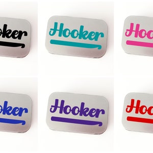May include: Six small silver tins with the word "Hooker" printed in a stylized font on the front. Each tin has a different colored crochet hook graphic underneath the word. The colors are black, teal, pink, blue, purple, and red.