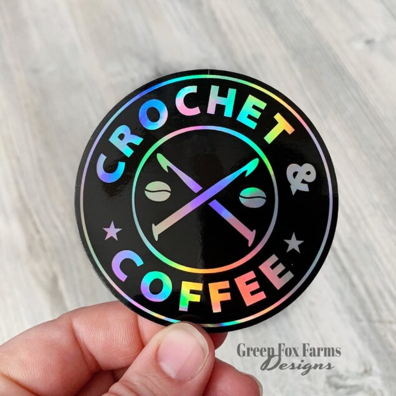 May include: A round, iridescent sticker with the text "Crochet & Coffee" in a black circle with a white star on each side. Two crossed crochet hooks and two coffee beans are in the center of the circle.