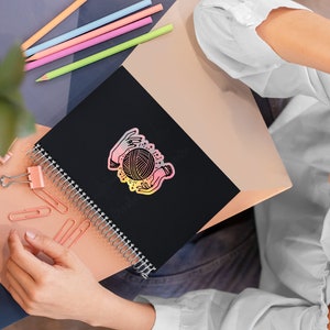 May include: A black spiral bound notebook with a colorful sticker of two hands holding a ball of yarn. The sticker has a rainbow gradient effect.