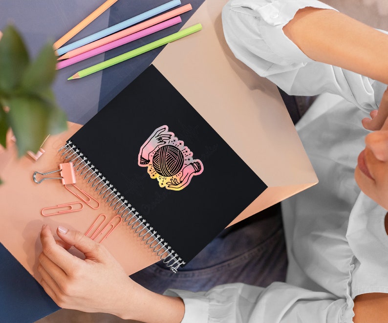 May include: A black spiral bound notebook with a colorful sticker of two hands holding a ball of yarn. The sticker has a rainbow gradient effect.