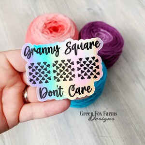 May include: A holographic sticker with a rainbow gradient background. The sticker features a black and white granny square design and the text "Granny Square Don't Care".