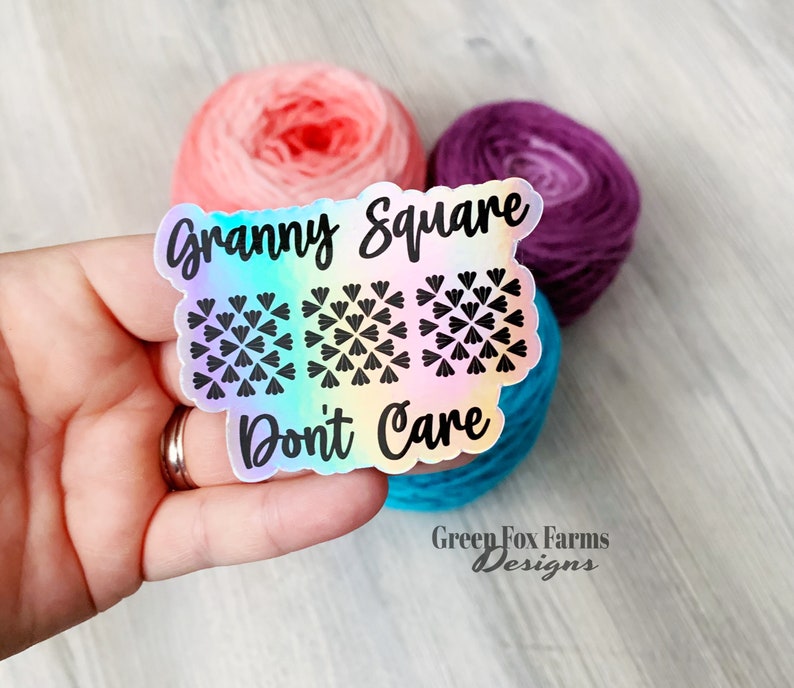 May include: A holographic sticker with a rainbow gradient background. The sticker features a black and white granny square design and the text "Granny Square Don't Care".