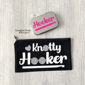 May include: Black canvas zippered pouch with the text "Knotty Hooker" in white with a white crochet hook and yarn balls. A silver tin with the text "Hooker" in pink letters is also shown.