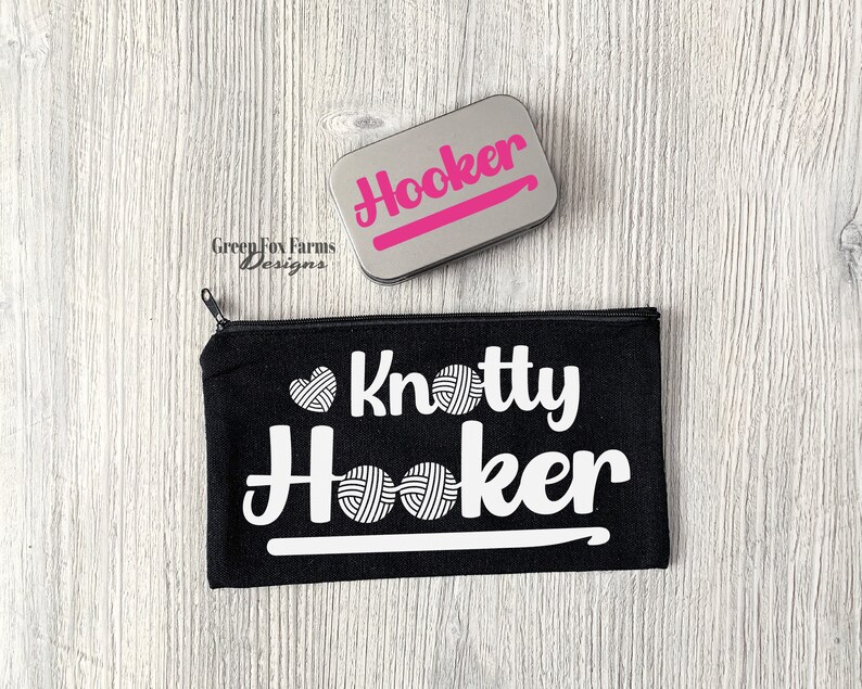 May include: Black canvas zippered pouch with the text "Knotty Hooker" in white with a white crochet hook and yarn balls. A silver tin with the text "Hooker" in pink letters is also shown.