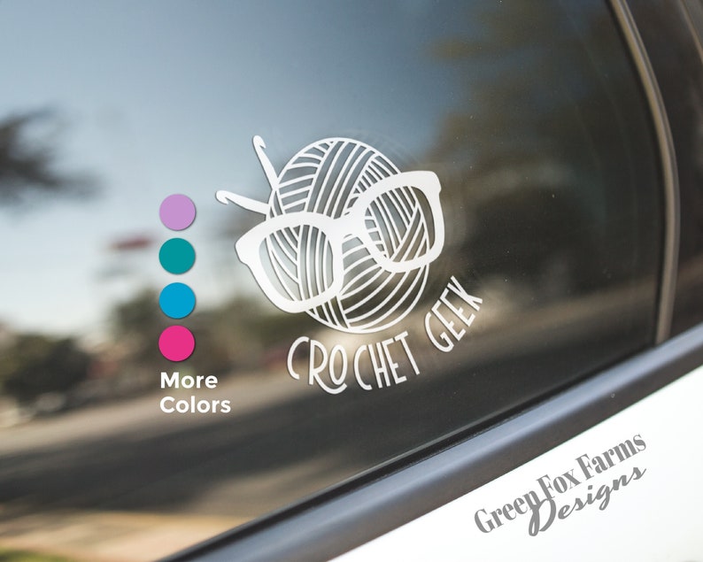 May include: White vinyl decal with a yarn ball wearing glasses and the text "Crochet Geek". The decal is on a car window.  The text "Green Fox Farms Designs" is also visible on the window.