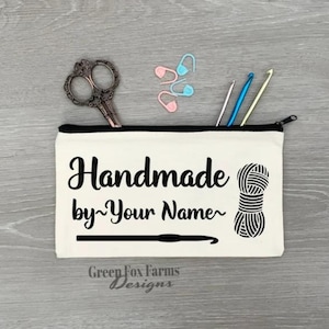 May include: A white canvas zippered pouch with the text "Handmade by~Your Name~" in black. The pouch also features a black and white illustration of a ball of yarn and a crochet hook. The text "Green Fox Farms Designs" is printed in black at the bottom of the pouch.