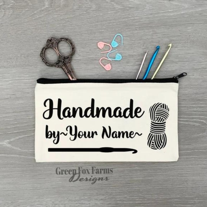 May include: A white canvas zippered pouch with the text "Handmade by~Your Name~" in black. The pouch also features a black and white illustration of a ball of yarn and a crochet hook. The text "Green Fox Farms Designs" is printed in black at the bottom of the pouch.