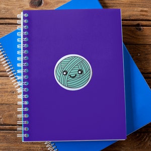 May include: A purple notebook with a spiral binding and a green yarn ball sticker with a smiling face. The notebook is on top of a blue notebook.