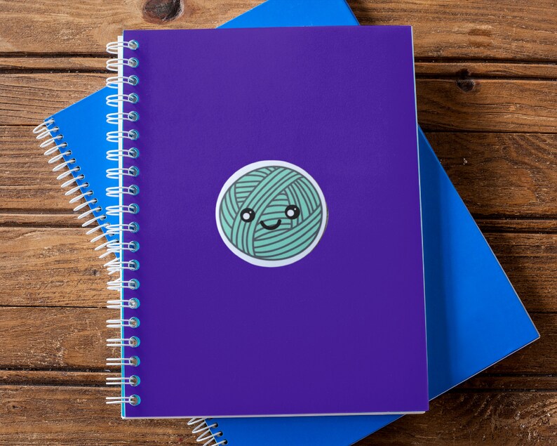 May include: A purple notebook with a spiral binding and a green yarn ball sticker with a smiling face. The notebook is on top of a blue notebook.