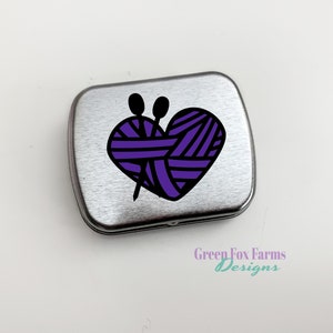 Puede incluir: Una lata de metal plateada con un dise&ntilde;o de ovillo de lana en forma de coraz&oacute;n morado y agujas de tejer. La lata tiene el texto "Green Fox Farms Designs" en la parte inferior.