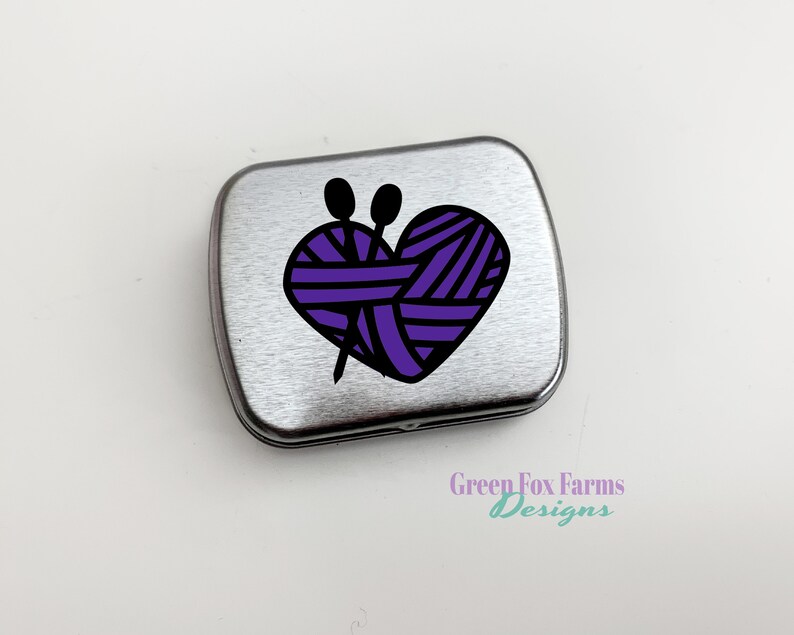 Puede incluir: Una lata de metal plateada con un dise&ntilde;o de ovillo de lana en forma de coraz&oacute;n morado y agujas de tejer. La lata tiene el texto "Green Fox Farms Designs" en la parte inferior.