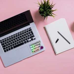 May include: A laptop computer with a sticker that reads "Introverted but willing to discuss YARN."  A notebook with a pen and a cup of coffee are on a pink surface.
